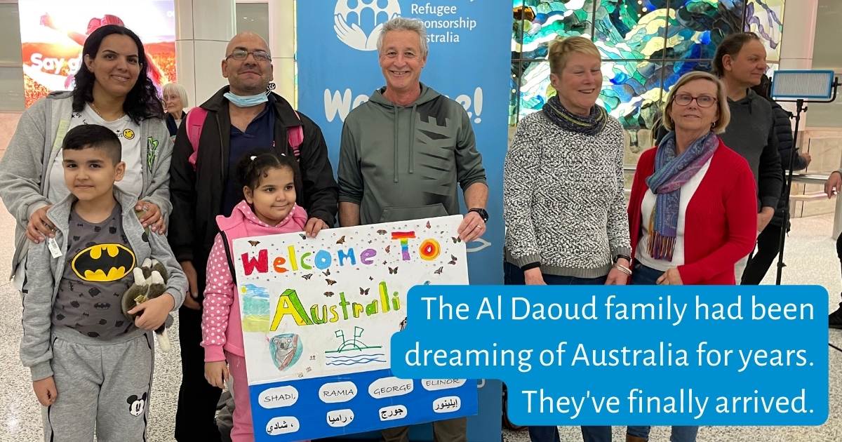 Family standing at airport, holding 'Welcome to Australia" sign, greeted by Community Supporter Group. "The Al Daoud family had been dreaming of Australia for years. They've finally arrived."